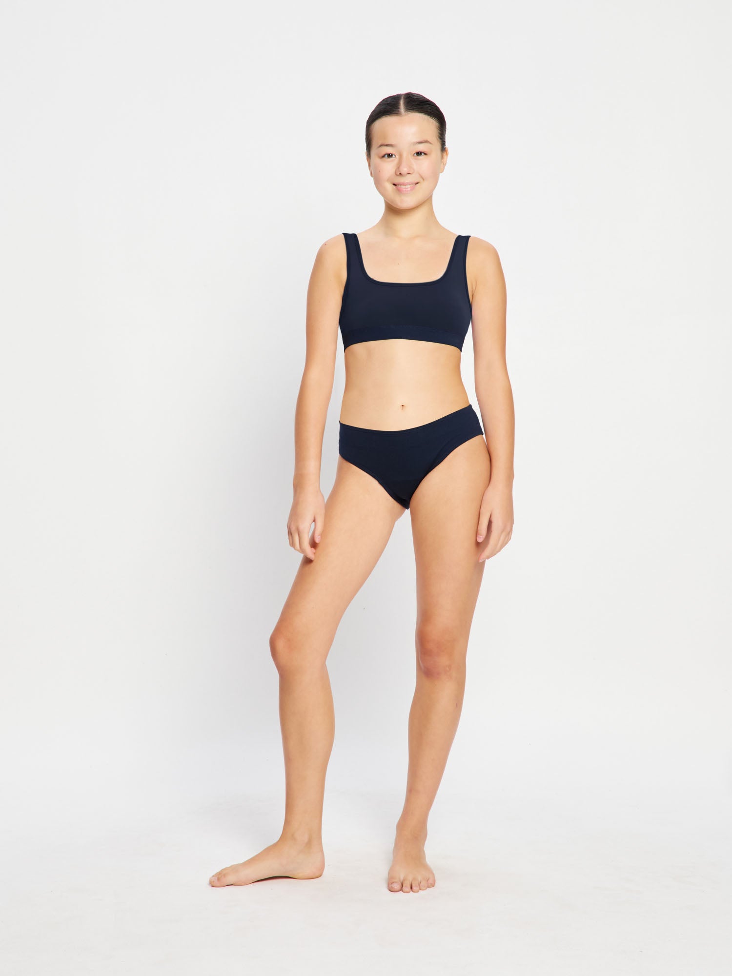 נערה בבגד ים ביקיני ספורטיבי שחור של מודיבודי, מבט מקדימה (NVMS), עומדת בביטחון על רקע חוף ים. מידת הדוגמנית - 10-12