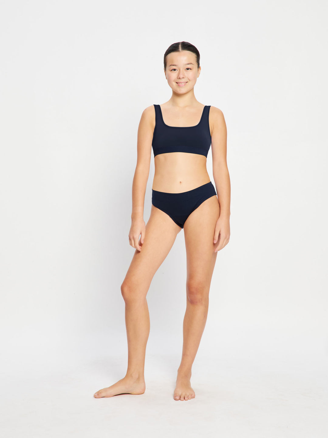 נערה בבגד ים ביקיני ספורטיבי שחור של מודיבודי, מבט מקדימה (NVMS), עומדת בביטחון על רקע חוף ים. מידת הדוגמנית - 10-12